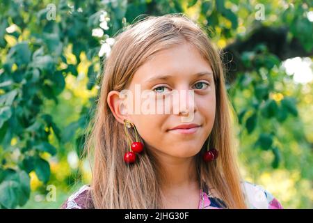 Defocus beautiful blond teen girl with sweet cherry on ear. Beautiful smiling teenage girl closeup, against green of summer park. Child earrings. Out Stock Photo