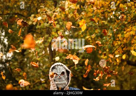 Defocus Halloween people. Person in grim reaper mask raising hand and throwing leaves. Many flying orange, yellow, green dry leaves. Skull ghost. Fall Stock Photo