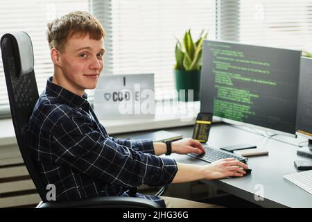 Portrait of young programmer looking at camera while sitting at his workplace with computer monitor and writing codes Stock Photo