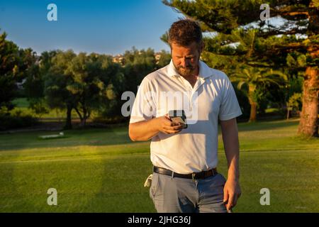 Portrait of young handsome golf player at golf club using smartphone. Stock Photo