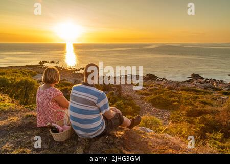 Married couple sitting on a cliff enjoing the sunset while having a picnic with wine and bread, Hovs hallar, Skåne , Sweden Stock Photo