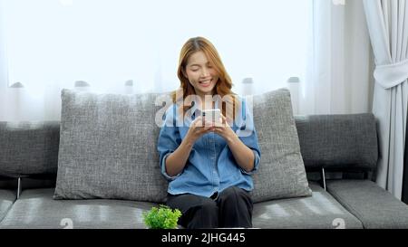 A smiling young woman using her phone and sitting on a couch Stock Photo
