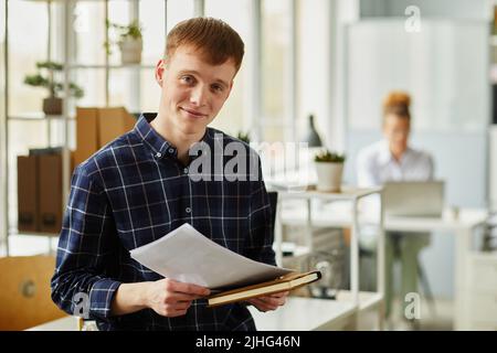 Portrait of young programmer in checked shirt holding notebook and documents and looking at camera while standing at office Stock Photo