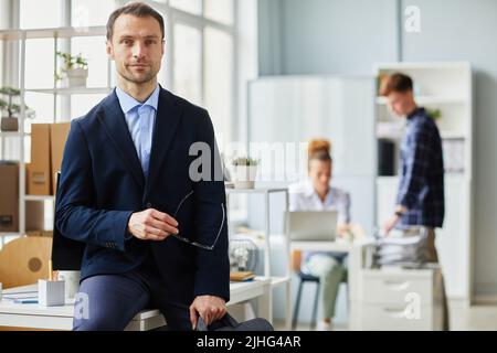 Portrait of mature professional programmer in jacket sitting on his workplace at office and looking at camera Stock Photo