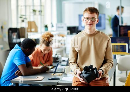 Portrait of young programmer in eyeglasses holding virtual reality glasses looking at camera while having meeting with his colleagues Stock Photo