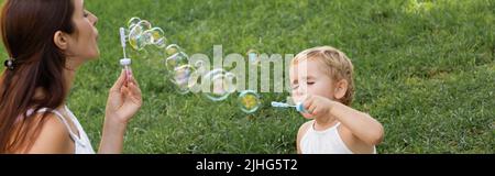 Mother and baby girl blowing soap bubbles in summer park, banner Stock Photo