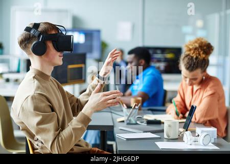 Young man in virtual reality glasses sitting at meeting with colleagues and testing new VR application Stock Photo