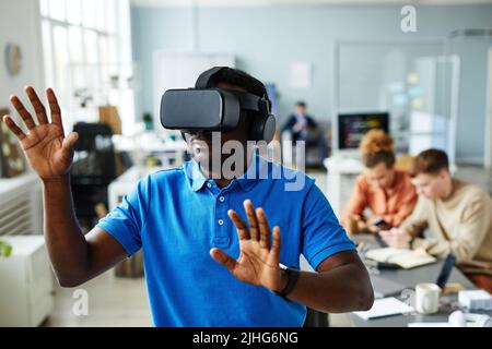 African young man in VR glasses gesturing while testing virtual reality program at office during meeting with colleagues Stock Photo
