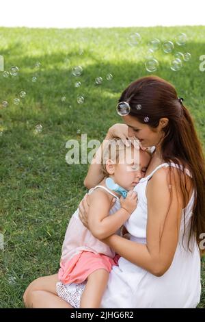 Smiling woman hugging toddler child near soap bubbles in park Stock Photo