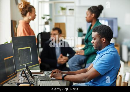 African young programmer writing codes on computer sitting at table with colleagues talking in background Stock Photo