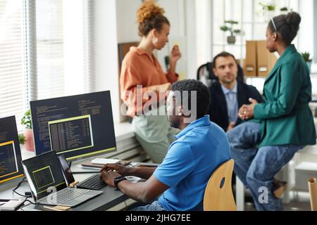 African IT specialist working with software typing on keyboard sitting at table with computers while his colleagues working in team in background Stock Photo