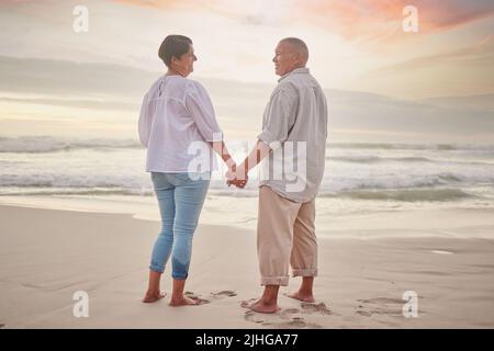 Rearview affectionate mature mixed race couple standing hand in hand on the beach. Senior husband and wife holding hands by the sea. They love Stock Photo