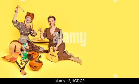 Women musicians in dresses with musical instruments on a yellow studio background. Happy artists with stringed musical instruments with smiles on face Stock Photo