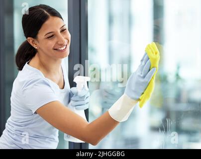 A happy smiling mixed race domestic worker using a cloth on a window. One Hispanic woman enjoying doing chores in her apartment Stock Photo