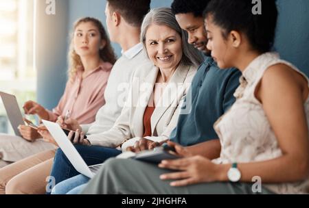 Group of happy businesspeople sitting on chairs in a row in an office together. Business professionals sitting and talking while working on a laptop Stock Photo
