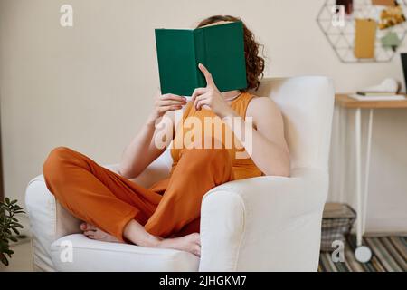 Young woman in orange casualwear reading book in dark green cover while sitting in white leather comfortable armchair in living room Stock Photo