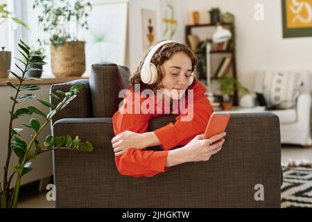 Cute adolescent girl in red shirt and white headphones looking at smartphone acreen while communicating in video chat Stock Photo