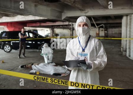 Young female crime scene investigator in protective coveralls making notes in document while standing against colleague over dead body Stock Photo