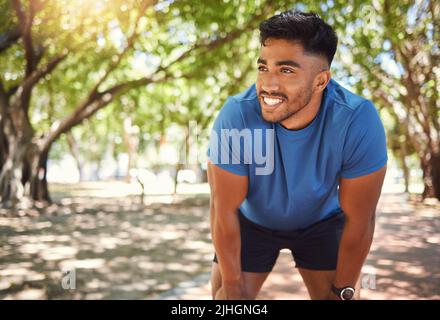 Happy athletic mixed race man smiling while standing with his hands on his knees in a park. Hispanic male resting after his run. Fit man pleased with Stock Photo