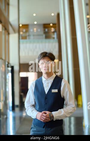 Young manager of hotel or bellboy in uniform standing in front of camera in corridor or lounge and waiting for new guests Stock Photo