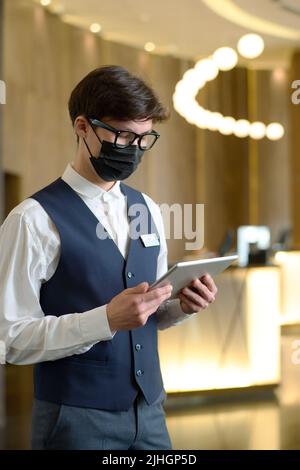 Male receptionist in uniform and protective mask checking online reservations of hotel rooms while standing in lounge Stock Photo