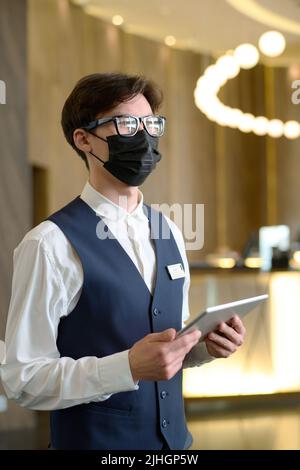 Young serious receptionist with tablet standing in spacious lounge of luxurious modern hotel while meeting new guests Stock Photo