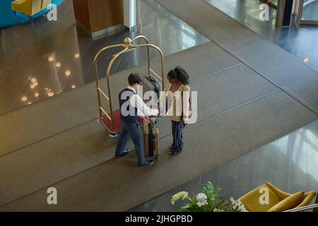 Above angle of young female traveler passing her suitcase to bellboy with cart standing in front of her in spacious lounge of hotel Stock Photo