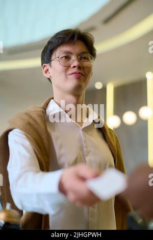 Asian guy in eyeglasses and white shirt passing keycard to receptionist by counter in lounge while going to leave hotel for work Stock Photo