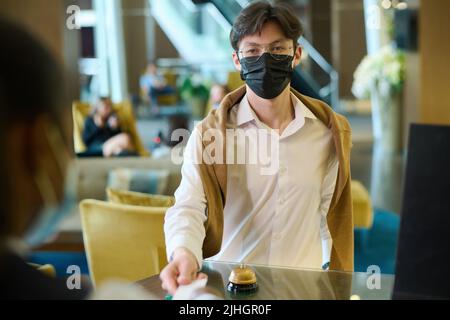 Young businessman in casualwear and black protective mask having registration by reception counter in lounge of modern hotel Stock Photo