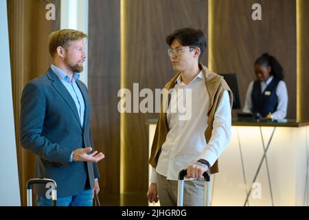 Two young intercultural businessmen with suitcase having discussion in lounge of modern hotel while standing against reception counter Stock Photo
