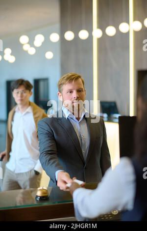 Confident businessman in formalwear taking keycard held by receptionist after registration in hotel while standing against his colleague Stock Photo