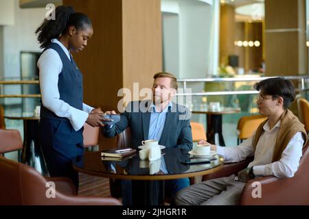 Young elegant businessman paying by credit card for two coffees in cafe while resting by table in the lounge of modern hotel after arrival Stock Photo