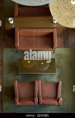 Above angle of brown leather sofa and two armchairs surrounding rectangular wooden table standing on grey carpet in hotel room Stock Photo