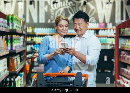 Happy couple of shoppers in supermarket, using phone to compare shopping list, man and woman shopping together Stock Photo