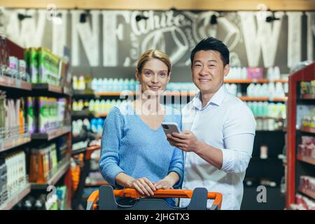 Family portrait of couple in supermarket shopping smiling and looking at camera, shoppers holding phone in hands, using discount app Stock Photo