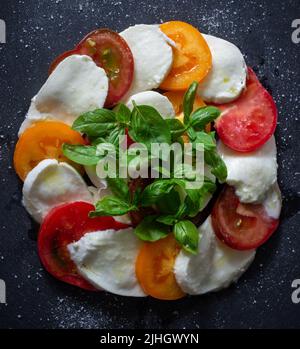 Fresh homemade caprese salad on a slate black plate. Mozzarella, basil and three kinds of tomatoes. Italian cuisine. Appetizer. Top view. Flat lay. Stock Photo