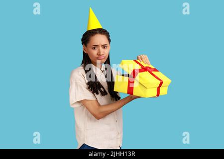 Portrait of unhappy upset woman in yellow party cone looking into gift box, opening present and looking at camera with sadness, wearing white shirt. Indoor studio shot isolated on blue background. Stock Photo