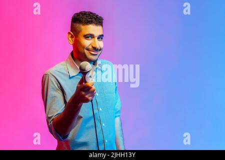 Portrait of man in shirt posing with microphone in hands, offers mic, journalist asking questions, looking smiling at camera. Indoor studio shot isolated on colorful neon light background. Stock Photo