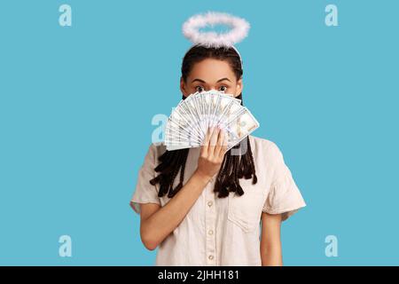 Angelic happy rich woman with black dreadlocks and nimbus on head peeking out of dollar banknotes, rejoicing big profit, wearing white shirt. Indoor studio shot isolated on blue background. Stock Photo