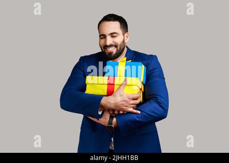 Satisfied bearded man with toothy smile embracing stack of present boxes, keeping eyes closed, enjoying holiday, wearing official style suit. Indoor studio shot isolated on gray background. Stock Photo