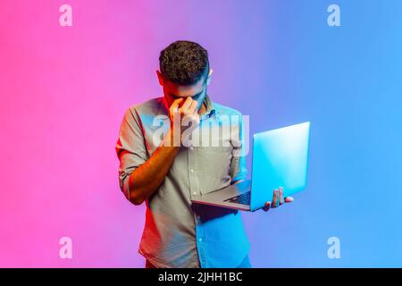 Portrait of tired man in shirt holding portable computer in hands and rubbing his eyes, being exhausted working long hours. Indoor studio shot isolated on colorful neon light background. Stock Photo