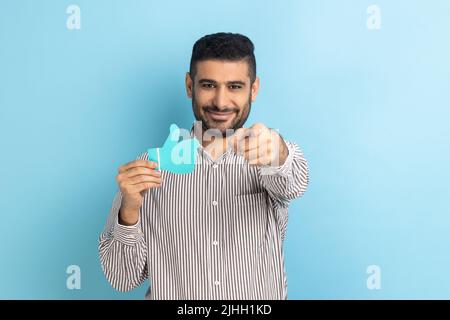Bearded businessman holding and showing like or thumbs up paper shape sign, pointing to camera with toothy smile, wearing striped shirt. Indoor studio shot isolated on blue background. Stock Photo