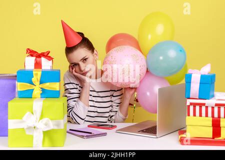 Depressed bored woman in party cone celebrating birthday sitting at workplace surrounded by many presents and air balloons. Indoor studio studio shot isolated on yellow background. Stock Photo