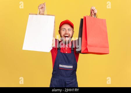 Portrait of delighted bearded courier man in blue uniform raised arms with shopping bags, satisfied with black friday big sale, bringing order. Indoor studio shot isolated on yellow background. Stock Photo