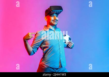 Happy smiling man in shirt wearing virtual reality glasses, standing with satisfied expression, pointing at himself with proud. Indoor studio shot isolated on colorful neon light background. Stock Photo