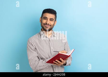 Delighted businessman with beard writing down to do list, making notes with pen in notebook, looking smiling at camera, wearing striped shirt. Indoor studio shot isolated on blue background. Stock Photo