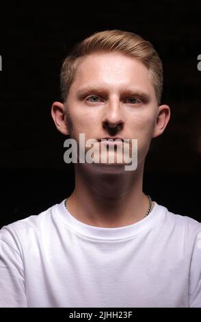 portrait of serious young man with folded and crossing hands standing on dark background. Guy in white t shirt is looking at camera. Confident concept Stock Photo