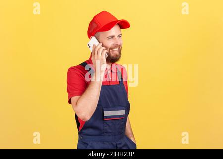 Portrait of happy courier or repairman in overalls taking phone with client, specify the time and place of completing work or delivery of goods. Indoor studio shot isolated on yellow background. Stock Photo