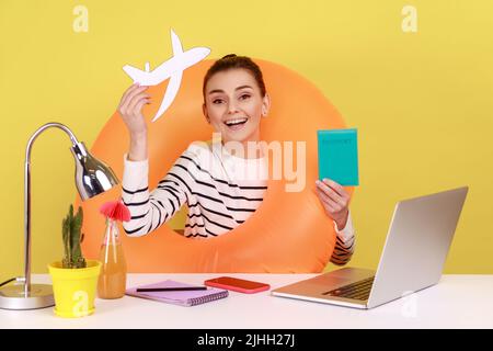 Extremely happy woman going on summer holidays, sitting at office with rubber ring and toy plane and showing passport, buying tickets. Indoor studio studio shot isolated on yellow background. Stock Photo