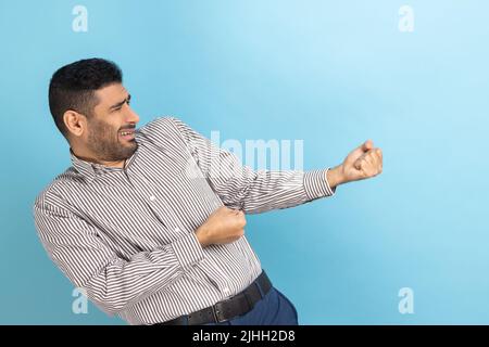 Side view of strong purposeful businessman with beard pulling invisible rope, showing his persistence and leadership qualities, wearing striped shirt. Indoor studio shot isolated on blue background. Stock Photo